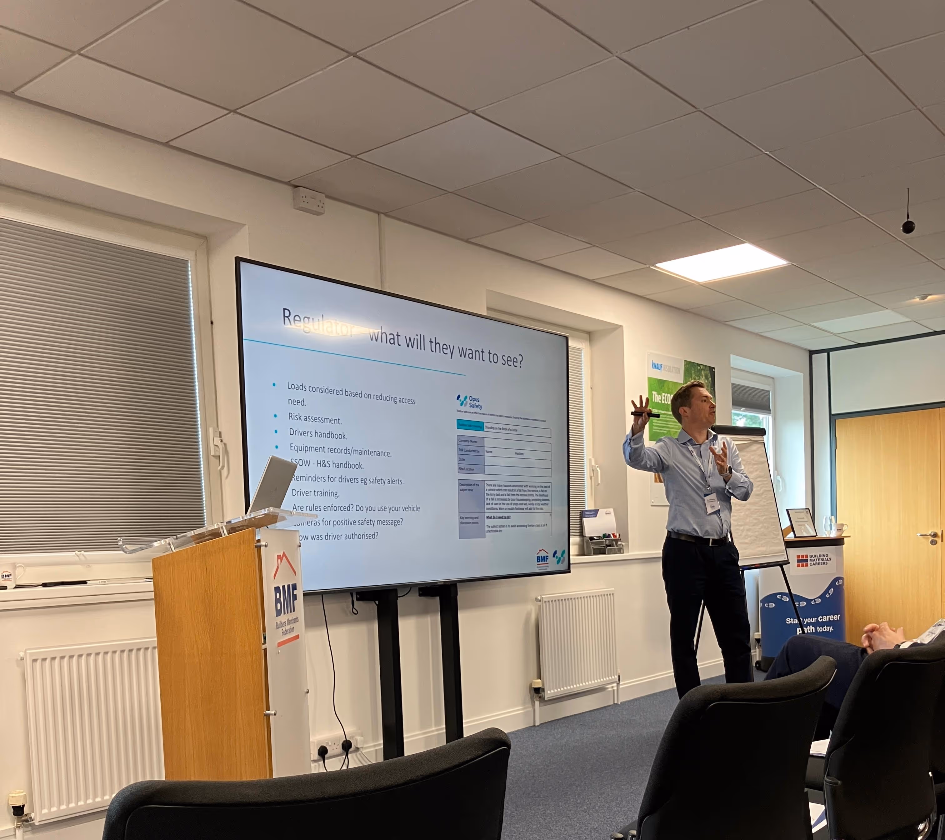 Man giving a presentation in a conference room with a screen displaying a slide titled 'Regulator what will they want to see?' and a podium labeled BMF Builders Merchants Federation.