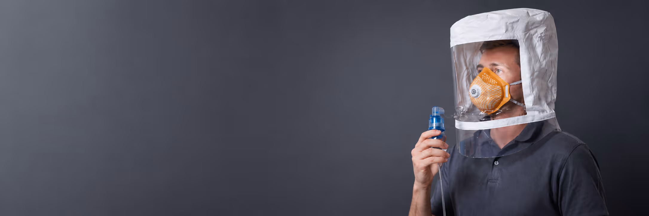 Man wearing an orange respirator mask and a clear protective hood holding a blue medical device against a gray background.