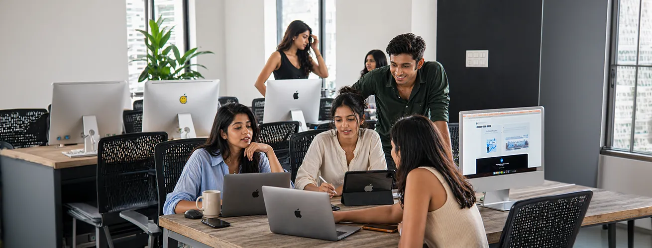 A group of five young professionals collaborating around laptops and a desktop in a modern office setting.