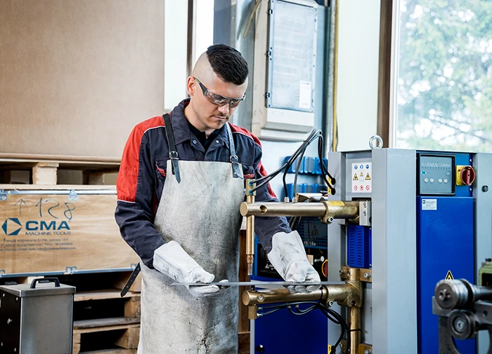 Mann schweißt Metallteile mit Handschuhen, Schutzbrille und Schürze an einer Schweißmaschine in Werkstatt.