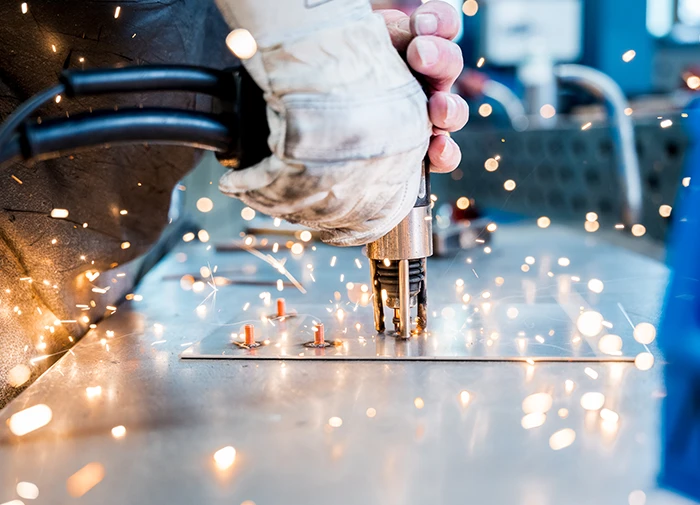 Person schweißt mit Handschuhen Funken fliegen beim Punktschweißen von Metallteilen.