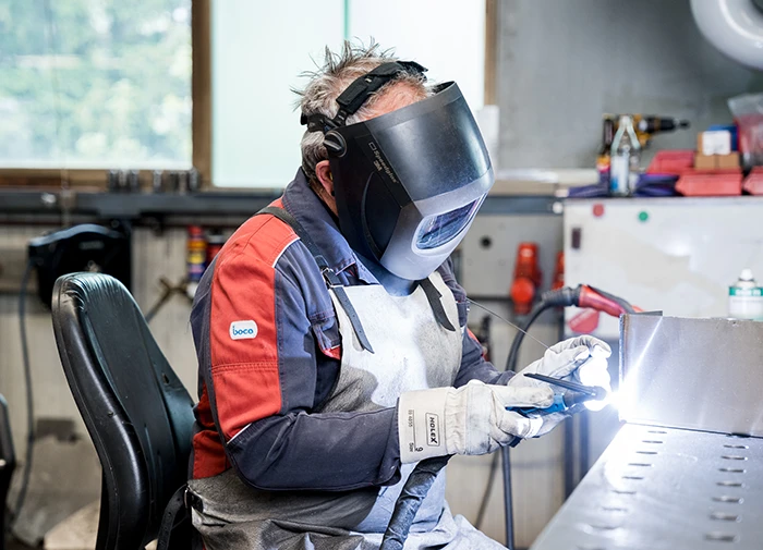 Handwerker beim Schweißen eines Metallteils mit Schutzschild, Handschuhen und Schweißgerät in Werkstatt.