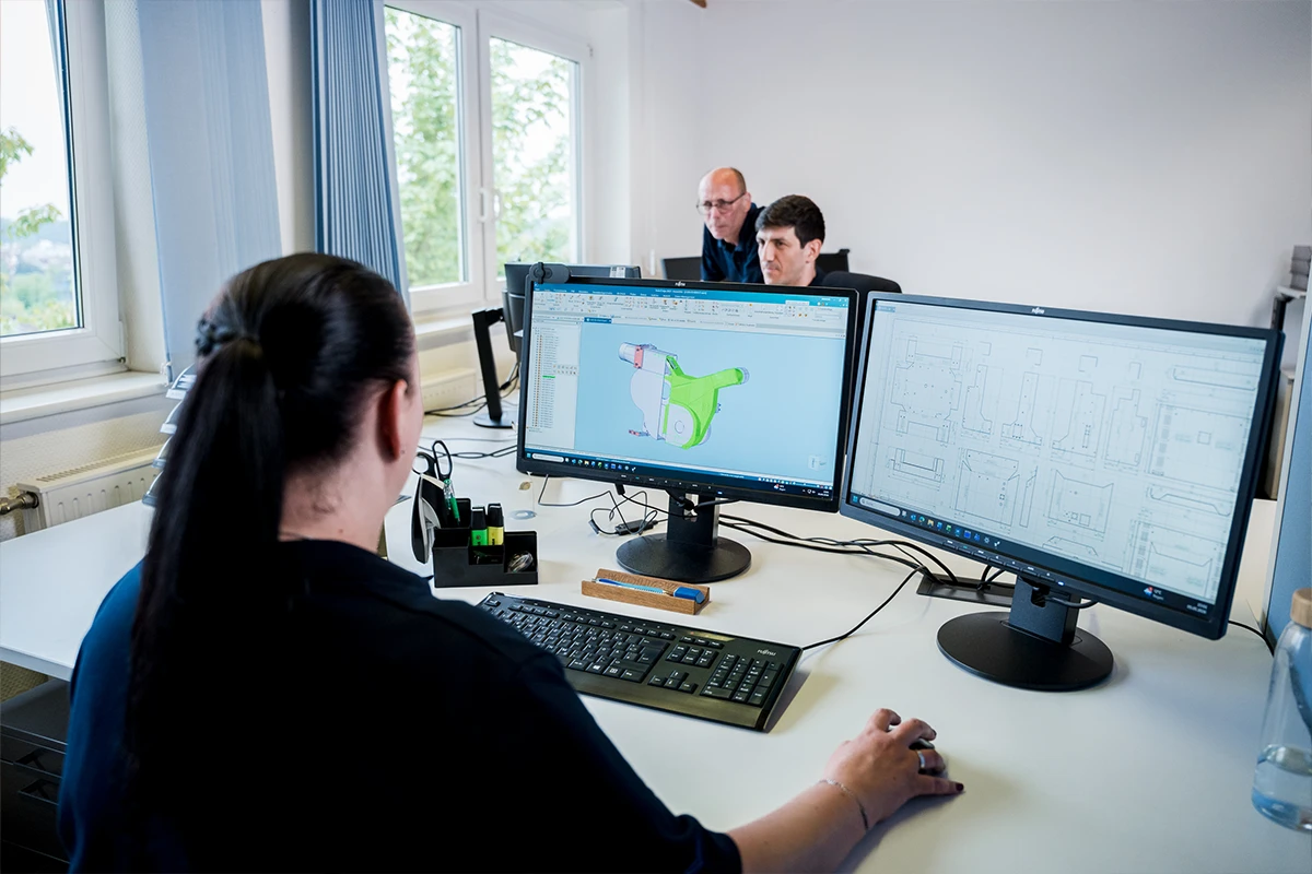Frau arbeitet an CAD-Konstruktion mit zwei Monitoren, zwei Männer im Hintergrund im Büro.