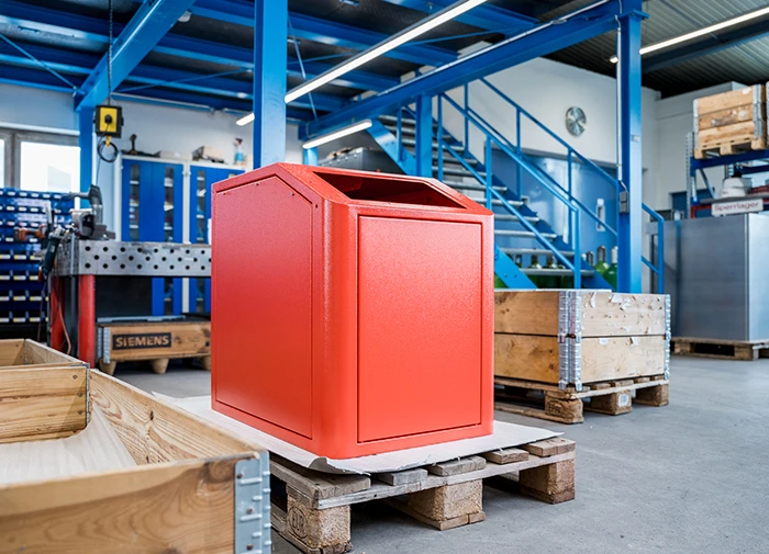 Roter Metallbehälter auf Holzpalette in industrieller Werkstatt mit blauen Stahlträgern und Regalen.