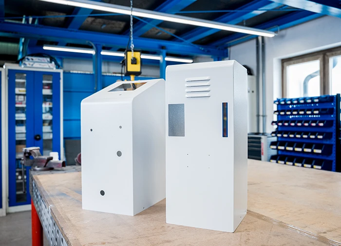 Zwei weiße lackierte Metallgehäuse stehen in einer Werkstatt auf einem Holztisch.