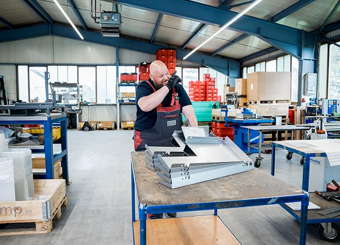 Arbeiter in Werkshalle telefoniert neben gestapelten Metallblechen auf einem Tisch.