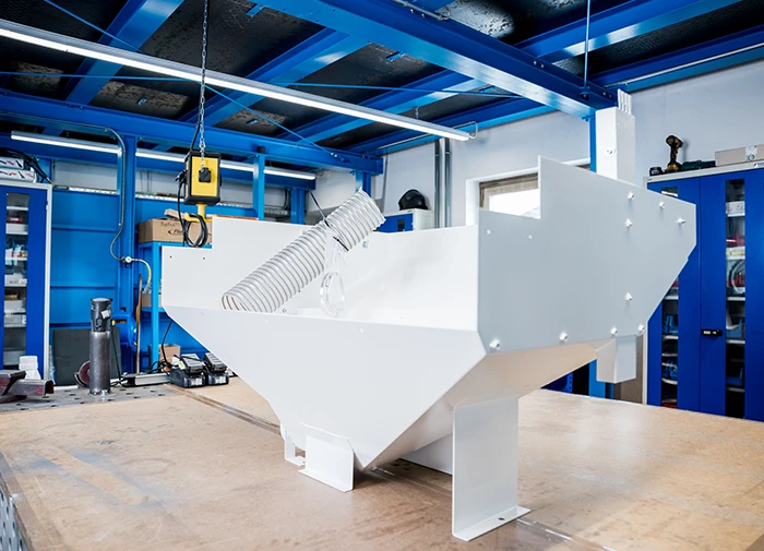 Weißes Metallteil mit Schlauch auf Tisch in Werkstatt mit blauen Regalen und Beleuchtung.