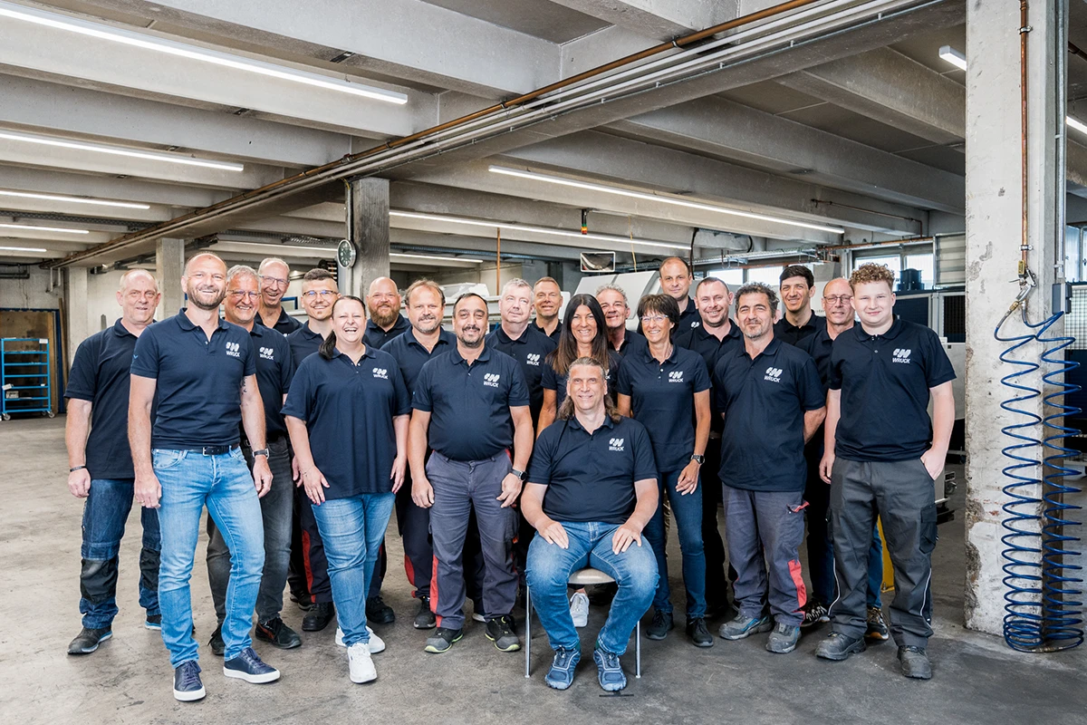 Gruppenfoto von 19 Mitarbeitern in Werkstattkleidung in einer Industriehalle.