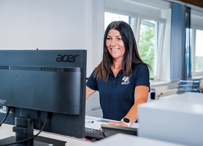 Frau mit dunkelblauem WRUCK-Poloshirt arbeitet lächelnd am Computer in hellem Büro.