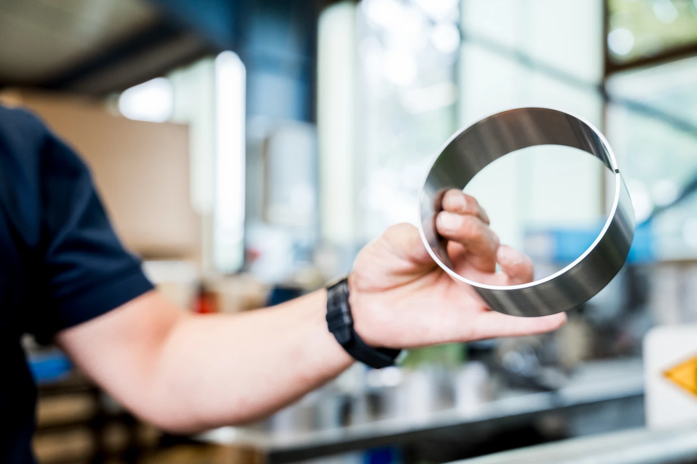 Hand hält glänzenden, runden Metallring in einer Werkstatt mit verschwommenem Hintergrund.