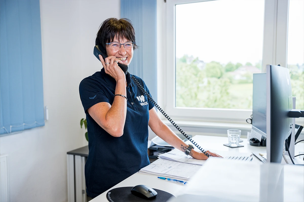 Frau mit Brille lächelt und telefoniert am Schreibtisch vor Computer und Fenster.