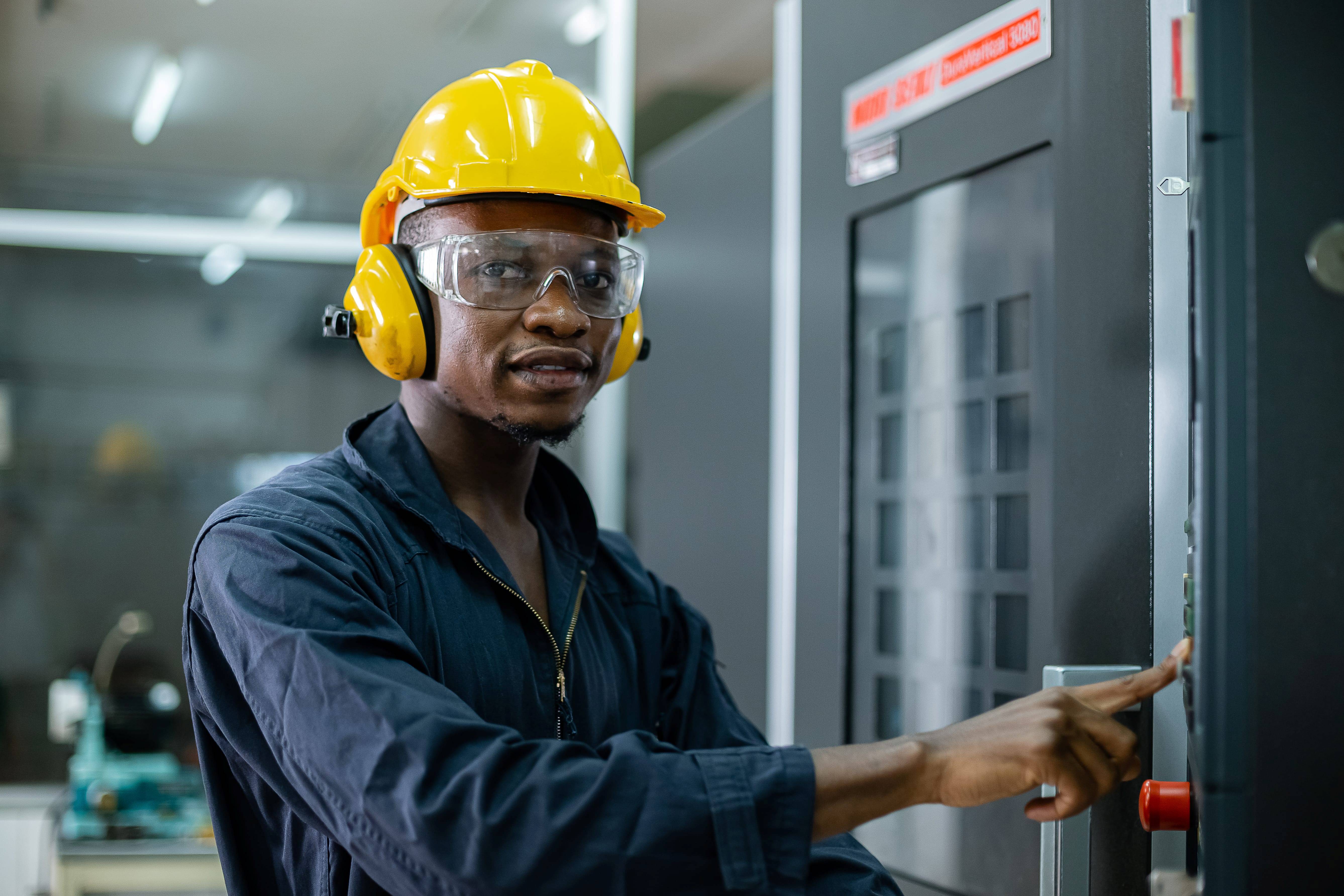 African American technician wearing soundproof headphones and a safety helmet while operating a CNC machine in an industrial factory