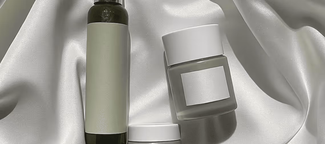 Three cosmetic containers with blank white labels on a light, silky fabric background.