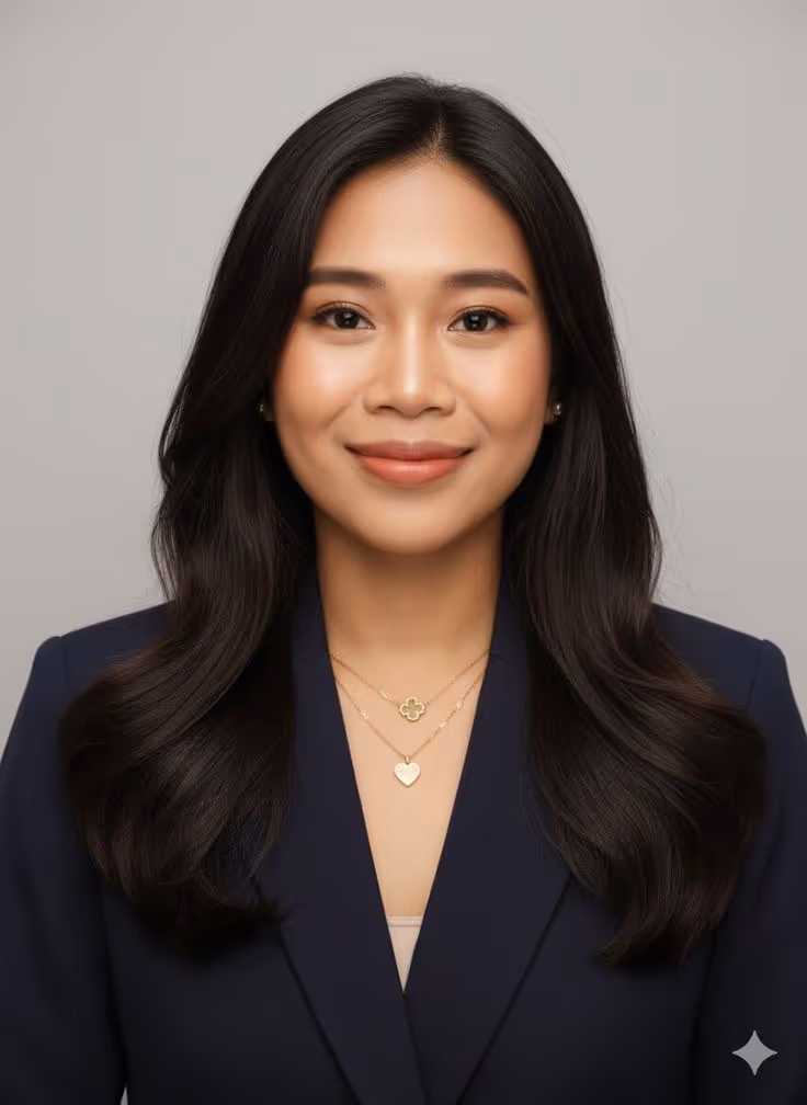 Portrait of a smiling woman with long black hair wearing a navy blazer and gold necklaces against a plain gray background.