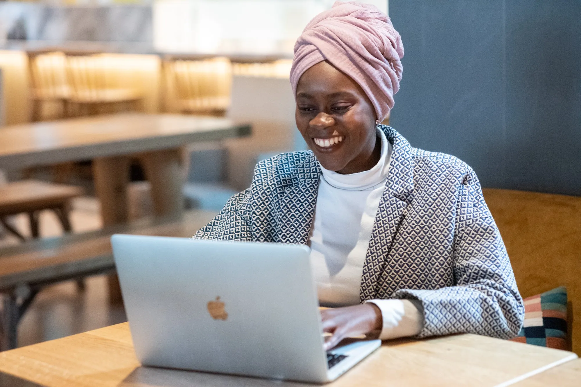 Smiling woman wearing a patterned jacket and pink headwrap working on a silver laptop at a wooden table in a cafe.