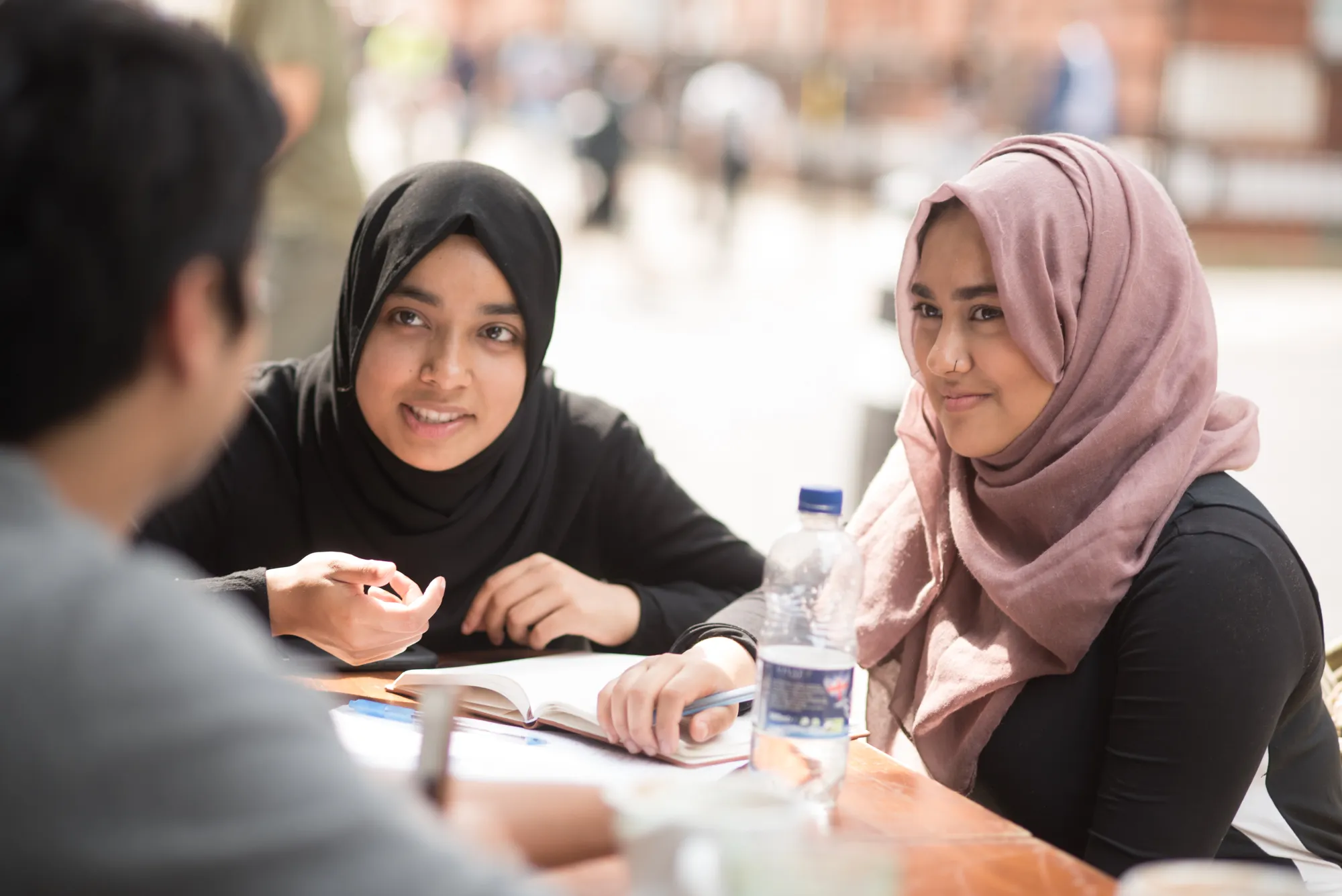 Smiling woman wearing a patterned jacket and pink headwrap working on a silver laptop at a wooden table in a cafe.