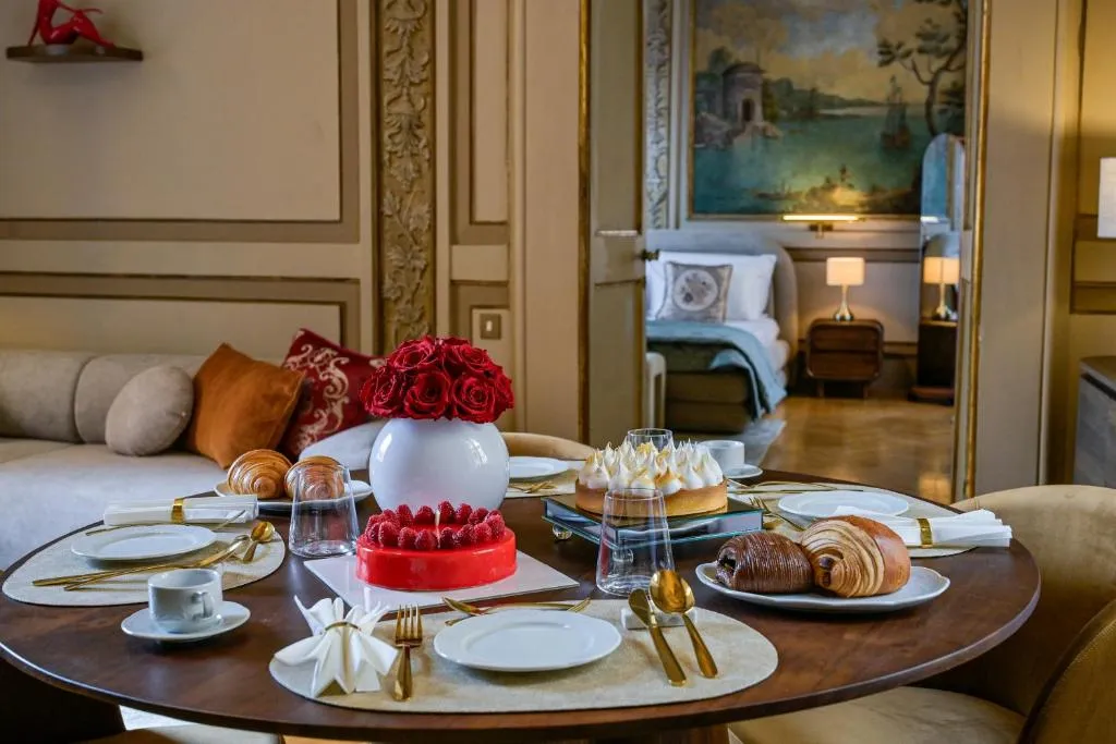 Round dining table set with plates, gold utensils, pastries, a raspberry cake, lemon meringue pie, a white vase with red roses, and a cozy living space and bedroom in the background.