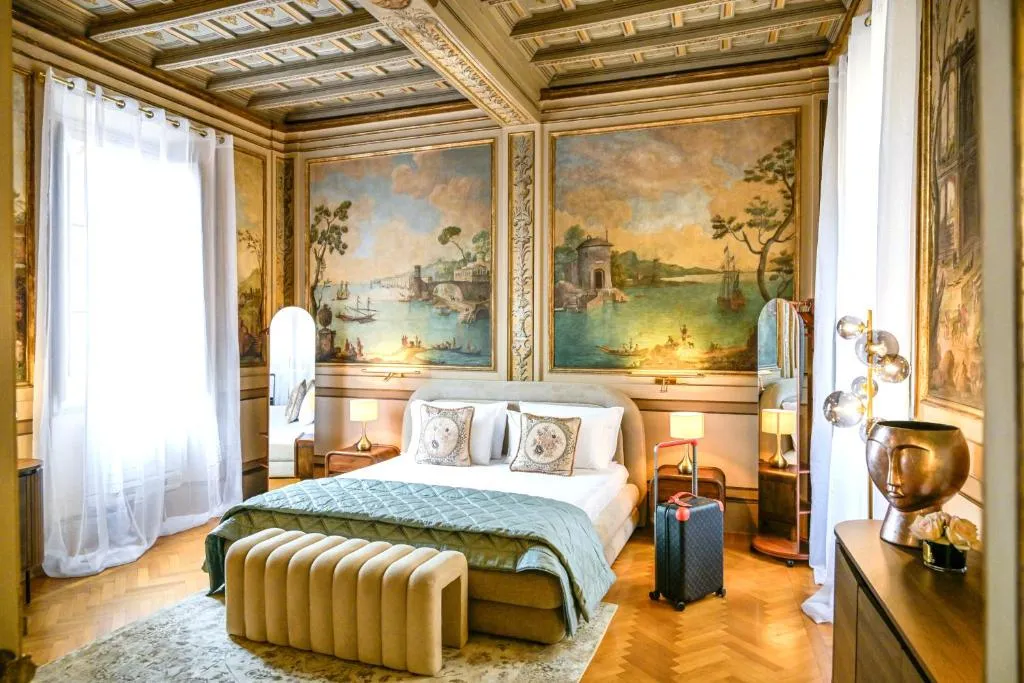 Luxurious bedroom with ornate ceiling, large landscape murals, sheer white curtains, a beige upholstered bed with patterned pillows, two standing mirrors, bedside lamps, and a modern gold decorative lamp on a wooden dresser.