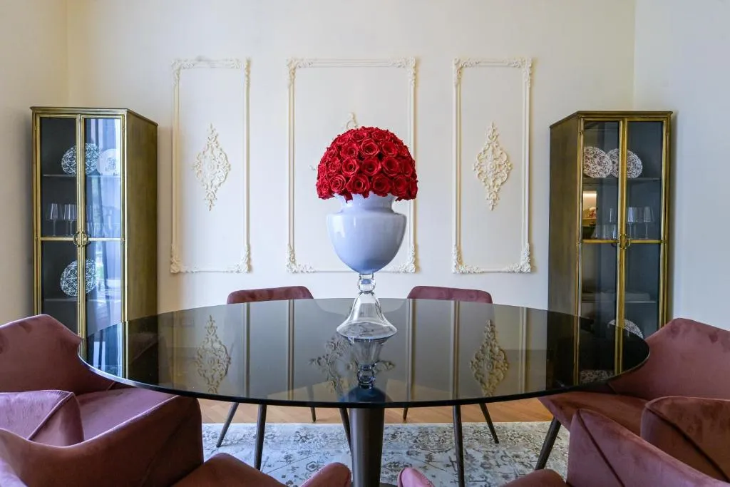 Elegant dining room with a round black glass table, pink velvet chairs, and a centerpiece of red roses in a white vase, flanked by two glass cabinets.
