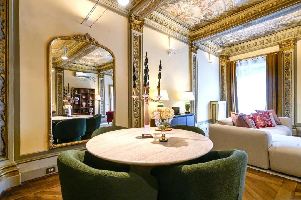 Elegant living room with ornate ceiling frescoes, gold-trimmed walls, a round table with green chairs, a large mirror, and a white sofa with decorative pillows.