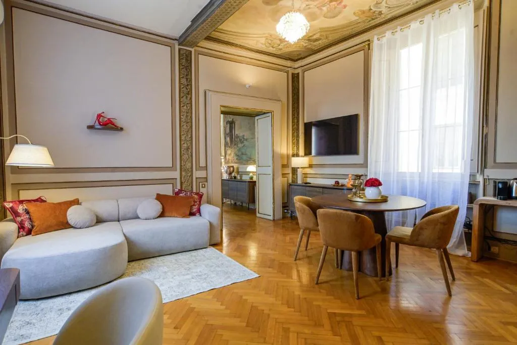 Elegant living room with a curved beige sofa, brown dining chairs around a round table, large window with sheer curtains, and detailed ceiling fresco.
