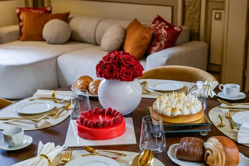 Dining table set with two cakes, croissants, red roses in a white vase, and place settings with gold cutlery and white napkins.