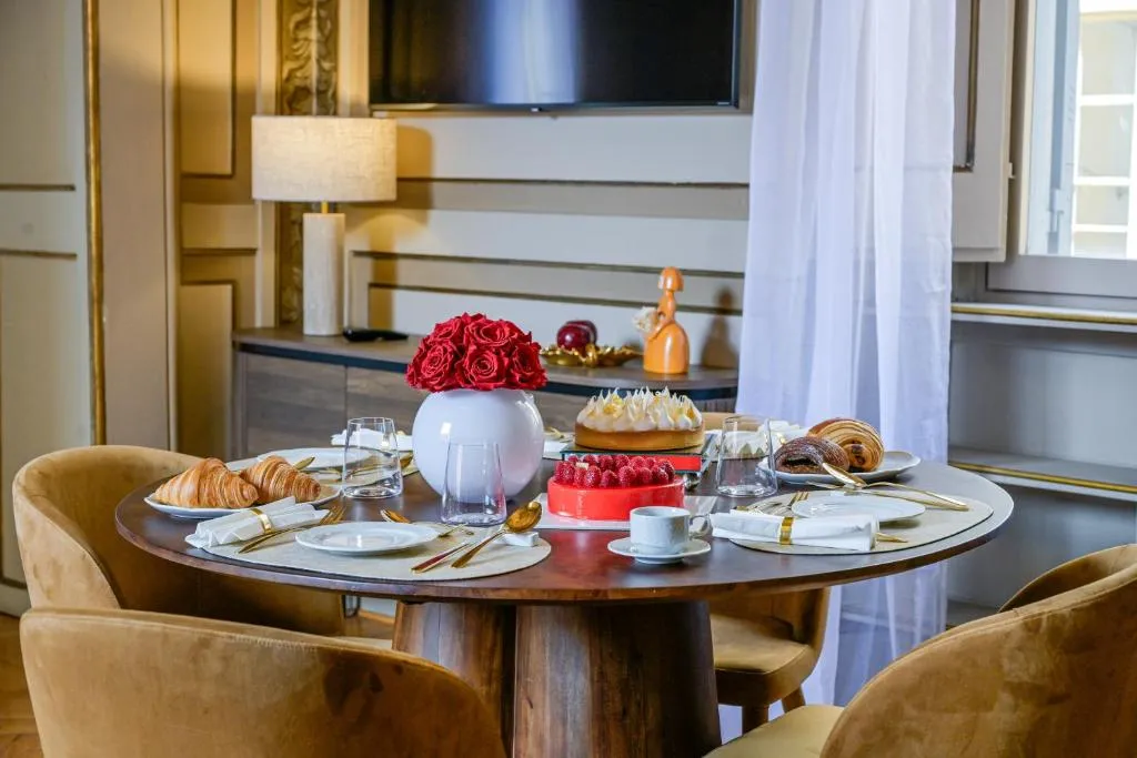 Round wooden dining table set for breakfast with croissants, cakes, coffee, glasses, and a vase of red roses, surrounded by brown chairs in a softly lit room.