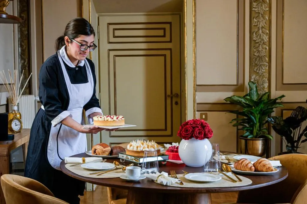 A woman in a maid uniform placing a dessert on a wooden dining table set with croissants, plates, cutlery, cups, and a vase of red roses.