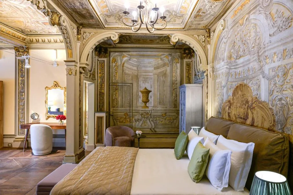 Luxurious bedroom with ornate classical ceiling and wall frescoes, large bed with green and white pillows, and a sitting area with armchairs.