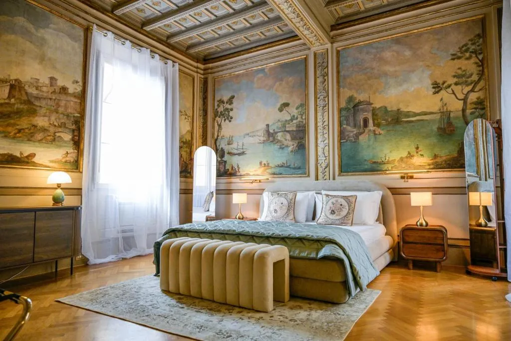 Luxurious bedroom with ornate ceiling and large wall murals depicting classical landscapes, featuring a bed with green bedding, side tables with lamps, and a cream-colored bench at the foot of the bed.