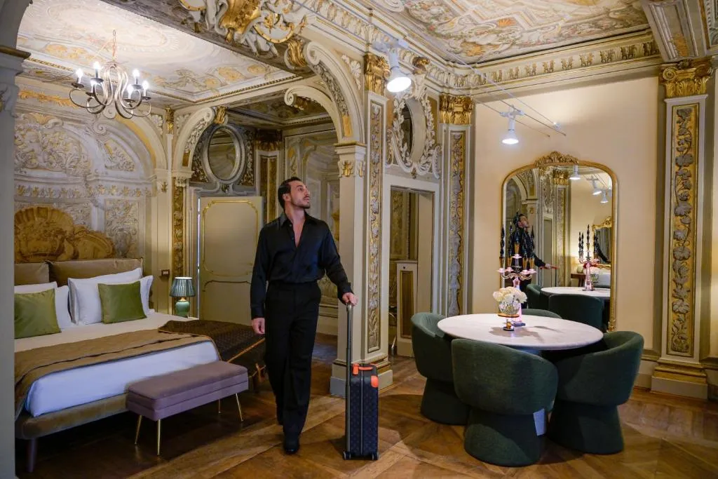 Man dressed in black standing with a suitcase in an ornately decorated room with a large bed, a round table with green chairs, and a large mirror reflecting the interior.