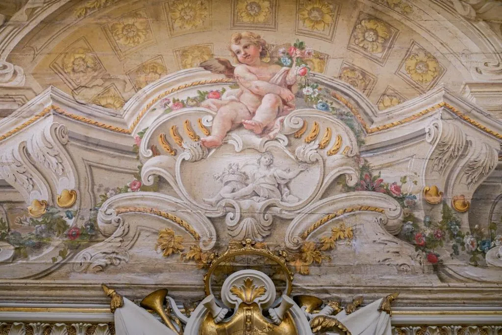Ornate ceiling fresco with a cherub holding flowers above intricate architectural details and floral decorations.
