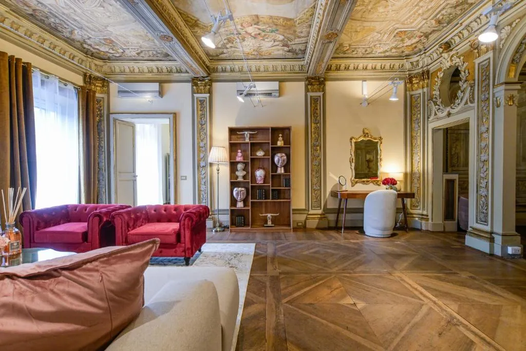 Luxurious room with ornate ceiling and walls, parquet wooden floor, red velvet armchairs, beige sofa, wooden bookshelf with decor, and a dressing table with a gold-framed mirror.