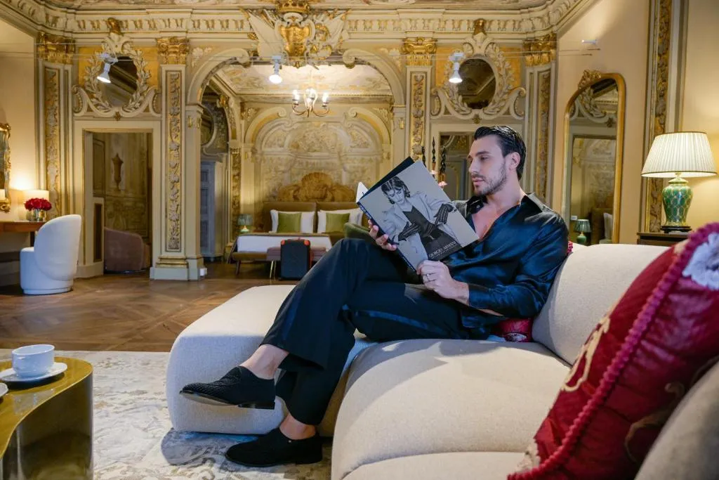 Man in black silk outfit sitting on cream sofa reading a fashion magazine in an ornately decorated room with gold accents.