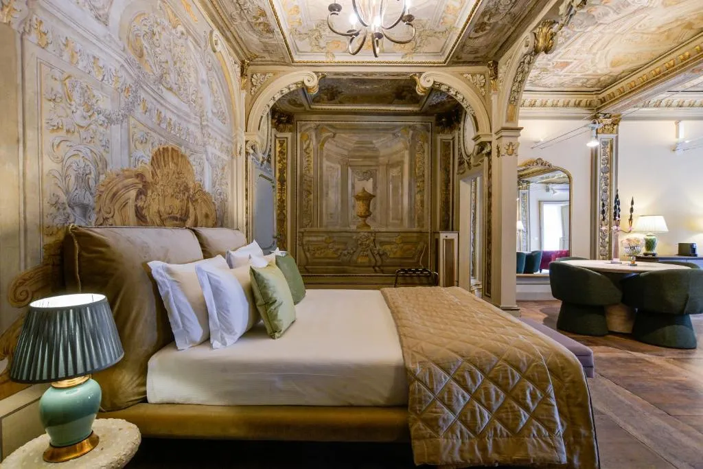 Luxurious bedroom with ornate frescoed walls and ceiling, a large bed with multiple pillows and a beige quilt, bedside lamp, and a seating area with green chairs and a mirror.