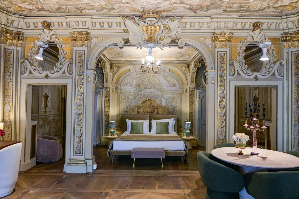 Luxurious hotel room with ornate ceiling and wall moldings, a bed with white linens and green pillows, bedside lamps, and a round dining table with green chairs.