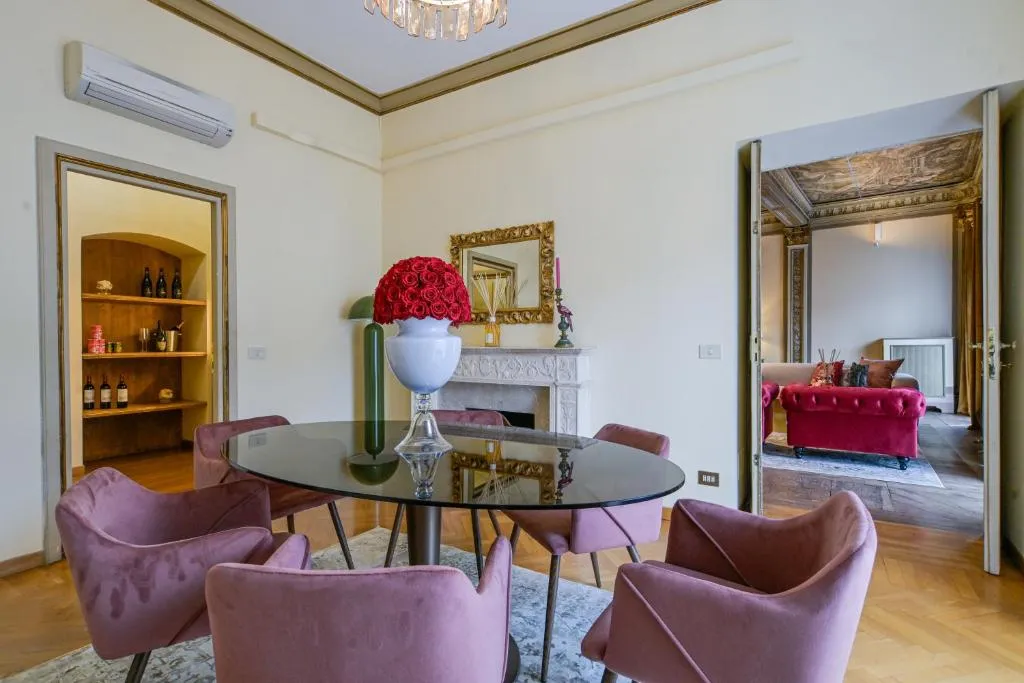 Dining room with a round glass table surrounded by six pink velvet chairs, a white vase with red roses on the table, and an open doorway leading to a room with a red velvet sofa.