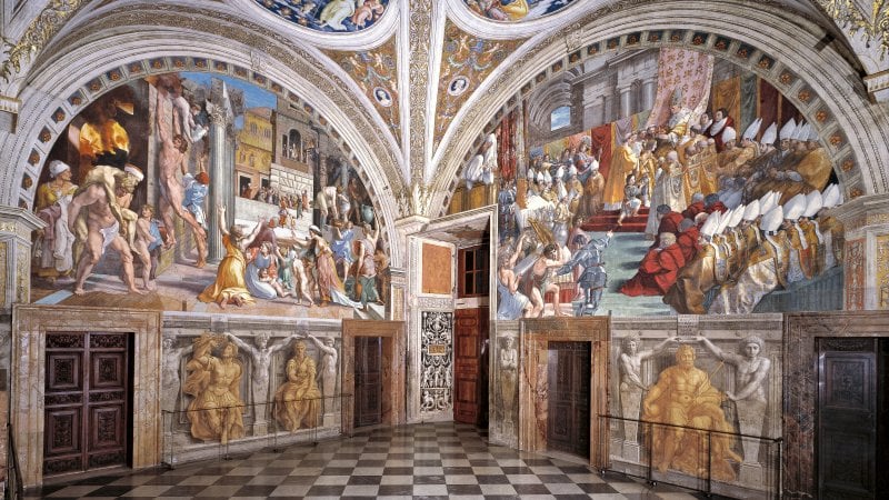 Interior view of a grand room with detailed Renaissance frescoes depicting various historical and religious scenes on arched walls and ceiling above a checkered floor.