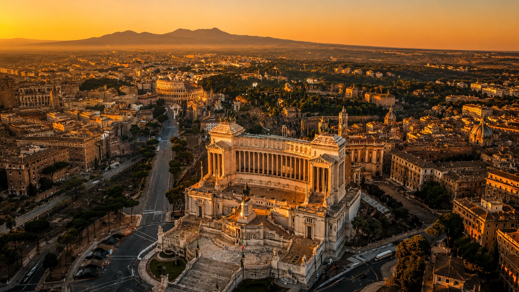 Aerial view of Rome at sunset with the Vittorio Emanuele II Monument in the foreground and the Colosseum visible in the background.