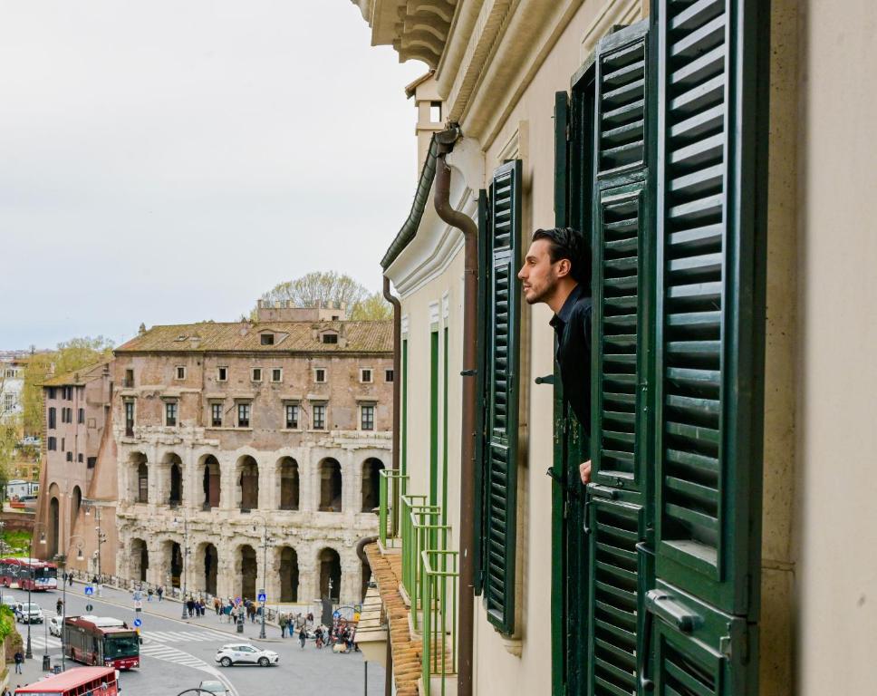 Man leaning out of window with green shutters overlooking ancient Roman ruins and a street with buses and pedestrians.