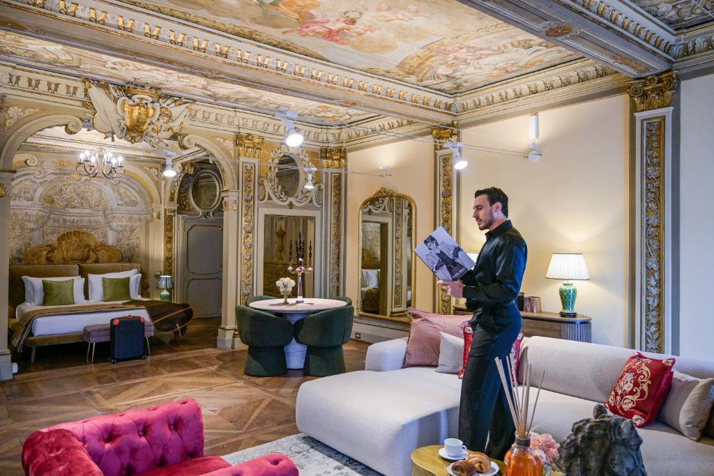 Man standing in an ornately decorated room with frescoed ceilings, reading a magazine near a beige sofa, with a bed and a round table in the background.