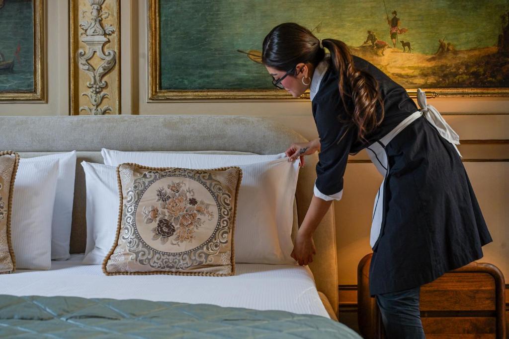 Hotel maid wearing glasses and a black uniform with a white apron adjusting a white pillow on a neatly made bed with decorative cushions.