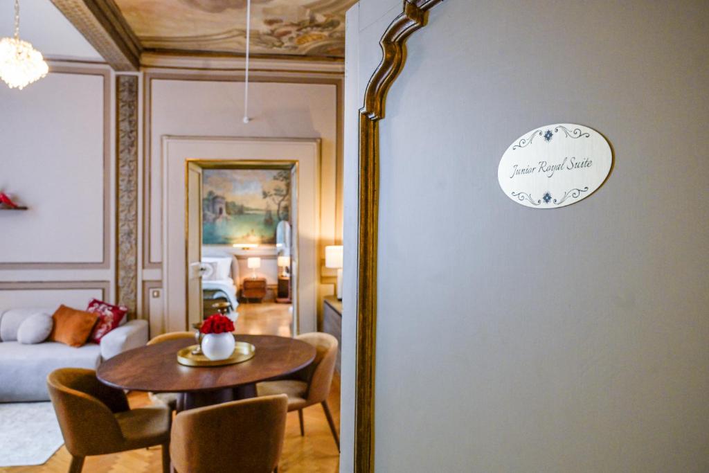 Interior of a Junior Royal Suite featuring a wooden dining table with four chairs and a white vase with red flowers, a sofa with red and orange cushions, and a bedroom visible through a doorway with a landscape mural on the wall.