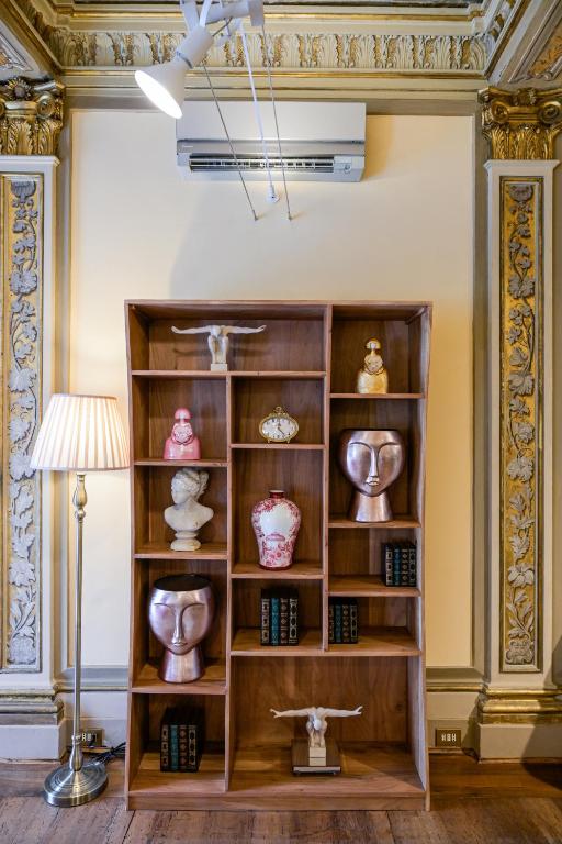 Wooden shelving unit with artistic busts, vases, sculptures, and vintage books, next to a tall floor lamp in a room with ornate gold-accented walls.