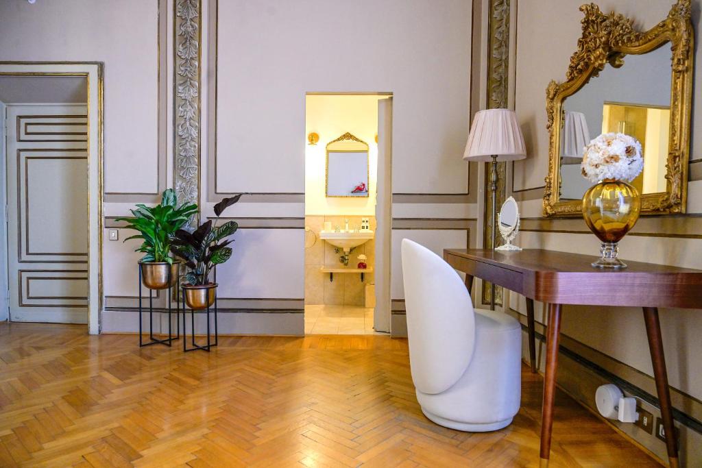 Elegant room with wooden parquet floor, ornate wall moldings, two potted plants, a white curved chair, a wooden table with a gold-framed mirror, lamp, and flower vase, and an open doorway leading to a bathroom.