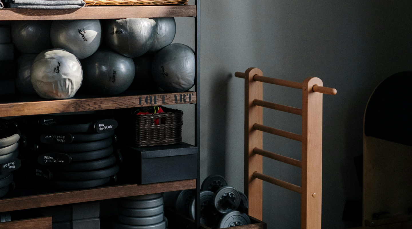 A cozy home gym corner featuring neatly stacked exercise balls, Pilates rings, and weights on wooden shelves beside a wooden ladder. Warm and organized ambiance.