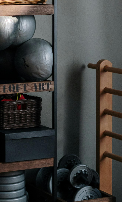 A cozy home gym corner featuring neatly stacked exercise balls, Pilates rings, and weights on wooden shelves beside a wooden ladder. Warm and organized ambiance.