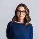 Smiling woman wearing glasses and a blue sweater against a light gray background.