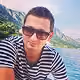 Man wearing sunglasses and a black and white striped shirt with a mountainous landscape and body of water in the background.