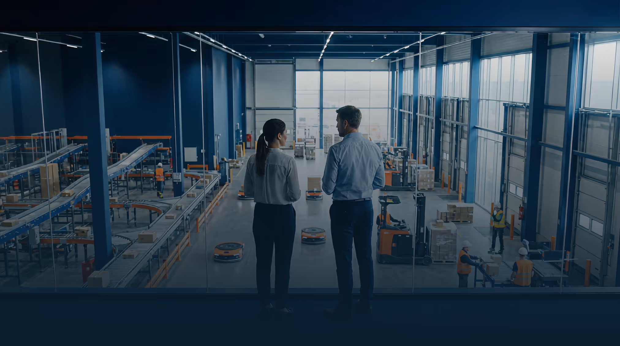 Twee personen kijken vanuit een glazen balkon uit op een magazijn met werkers, automatisering en transportbanden.